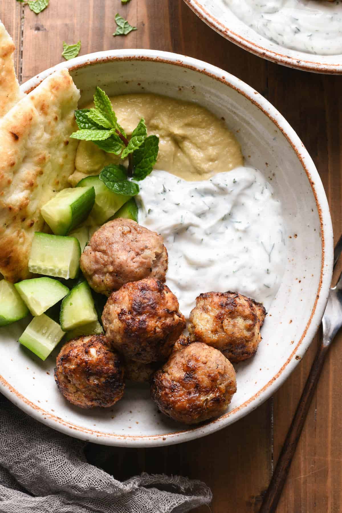 Are you looking for a turkey meatball that isn't dry? These Air Fryer Turkey Meatballs are for you! Cook in an air fryer or oven, and they turn out juicy and flavorful every time. | foxeslovelemons.com Overhead photo of rustic bowl filled with air fryer turkey meatballs, chopped cucumber, hummus, yogurt sauce, naan and a mint sprig garnish.