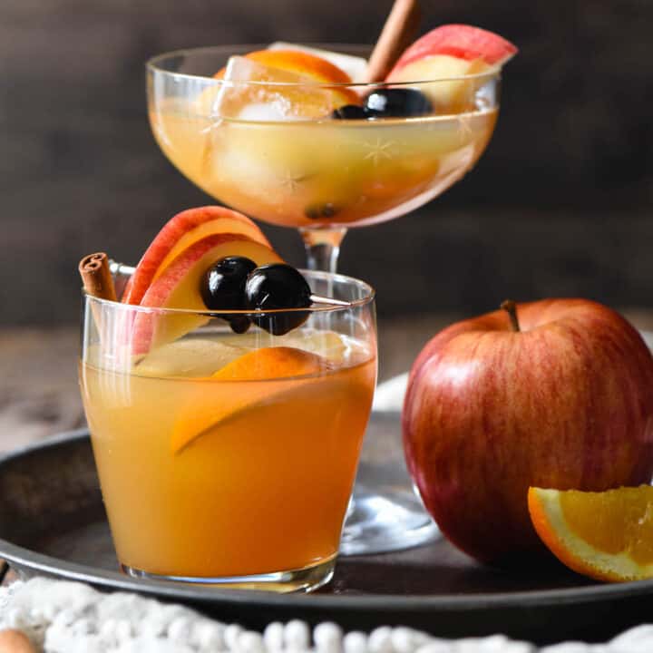 Apple cider old fashioned cocktails in two different glasses (1 tall, 1 short), on a tray with a whole apple.