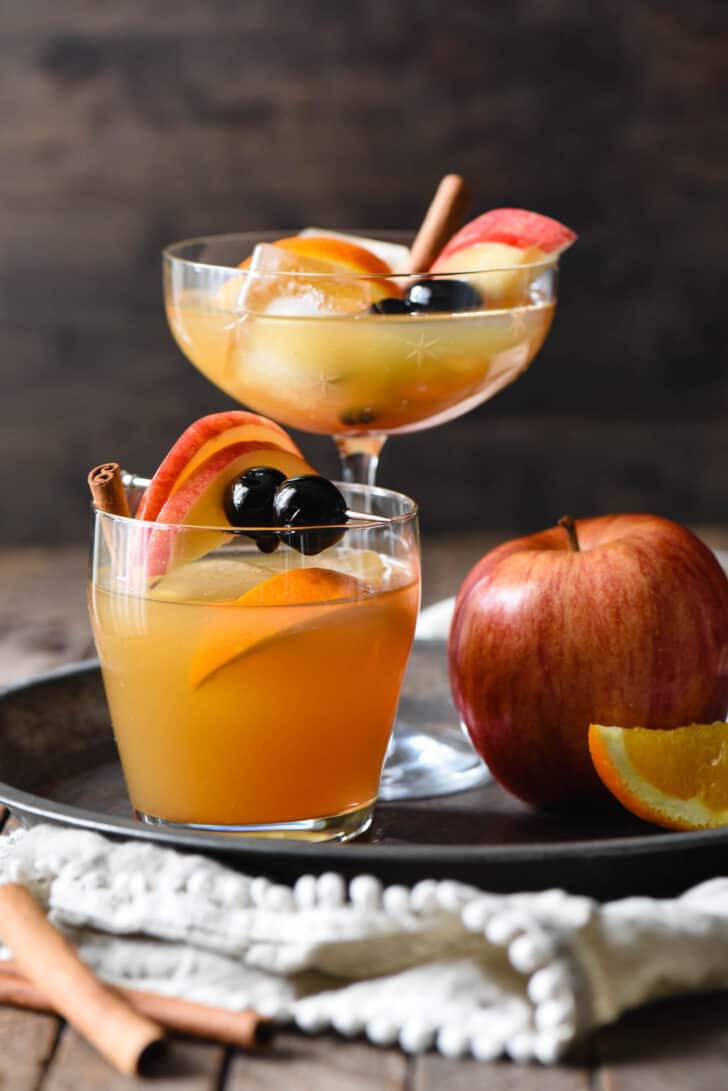 Apple cider old fashioned cocktails in two different glasses (1 tall, 1 short), on a tray with a whole apple.