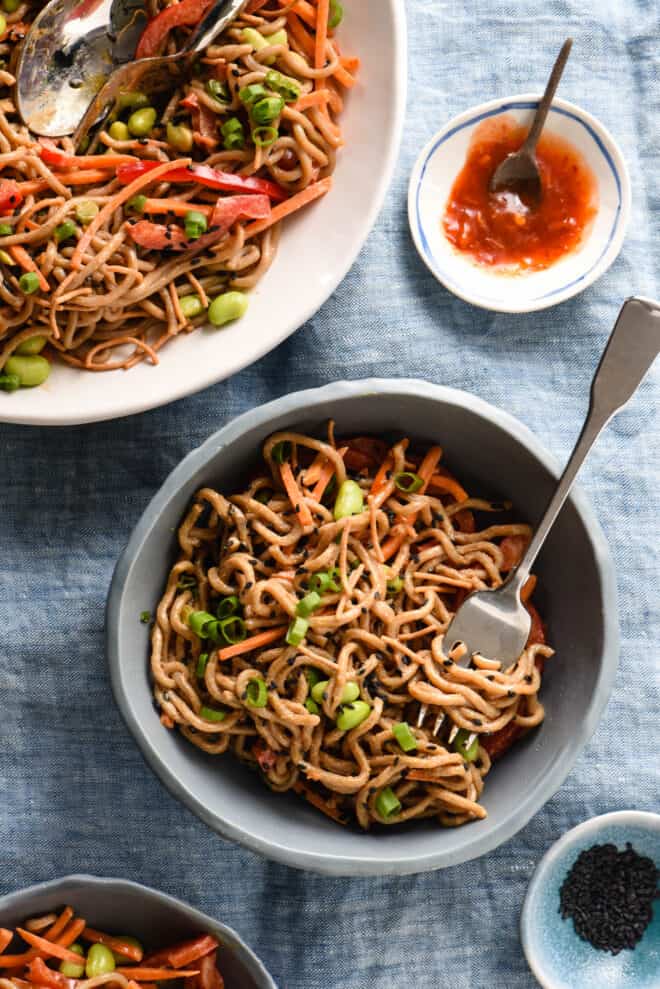 Slate blue bowl filled with Asian noodle salad, alongside larger serving bowl and small bowls of hot sauce and black sesame seeds.