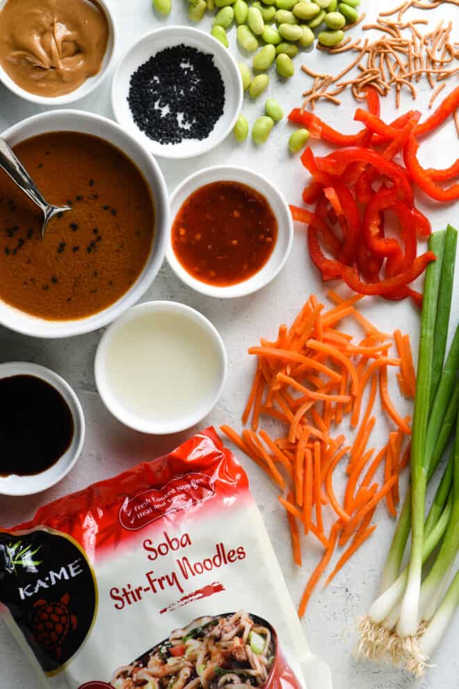 White tabletop filled with ingredients for cold noodle salad, including carrots, green onions, red pepper, edamame and Asian condiments.