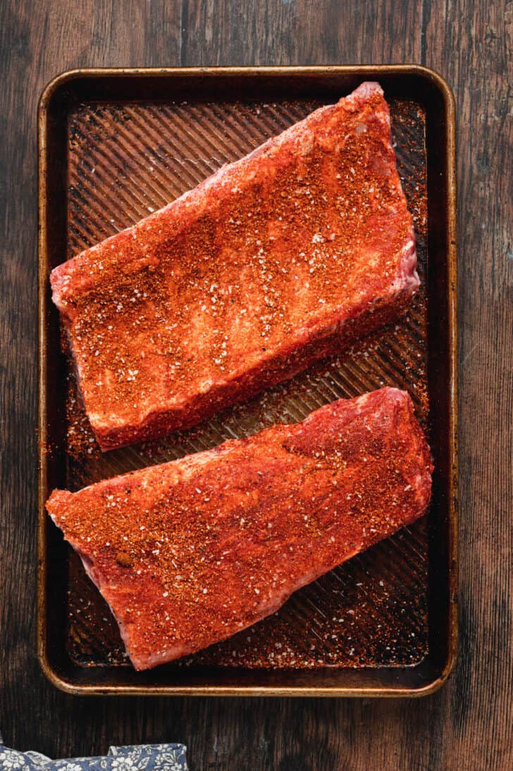 Baby back ribs rubbed with red spices on a textured baking pan.