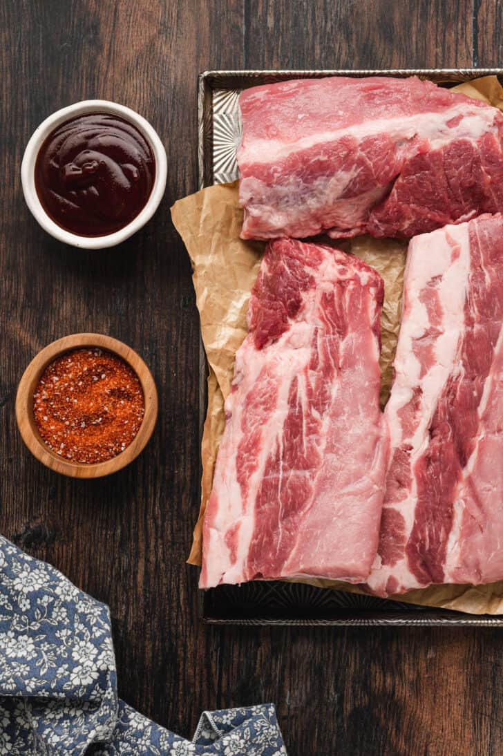 Ingredients laid out on a wooden surface, including bone-in pork, a red spice rub and barbecue sauce.