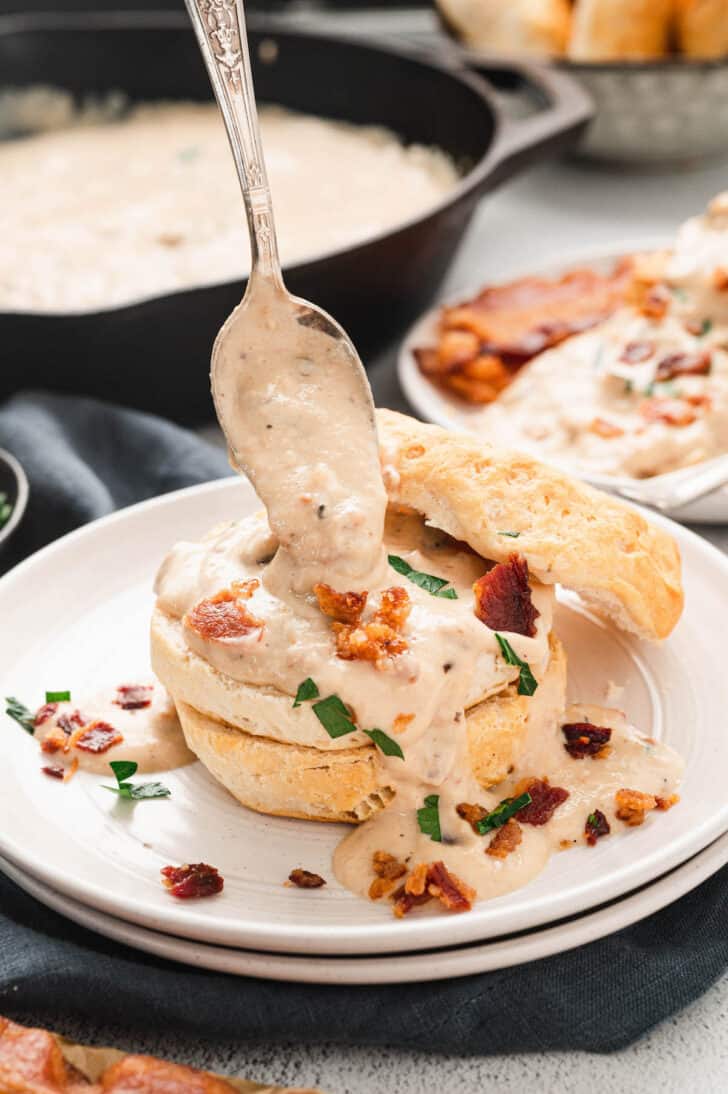 A biscuit cut in half on a small white plate, with bacon gravy being spooned over it.