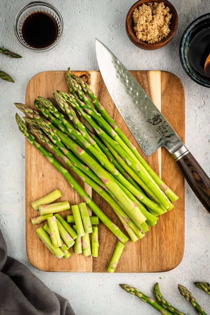 Fresh sparrow grass on a wooden cutting board in the process of being chopped, with a large chef's knife.