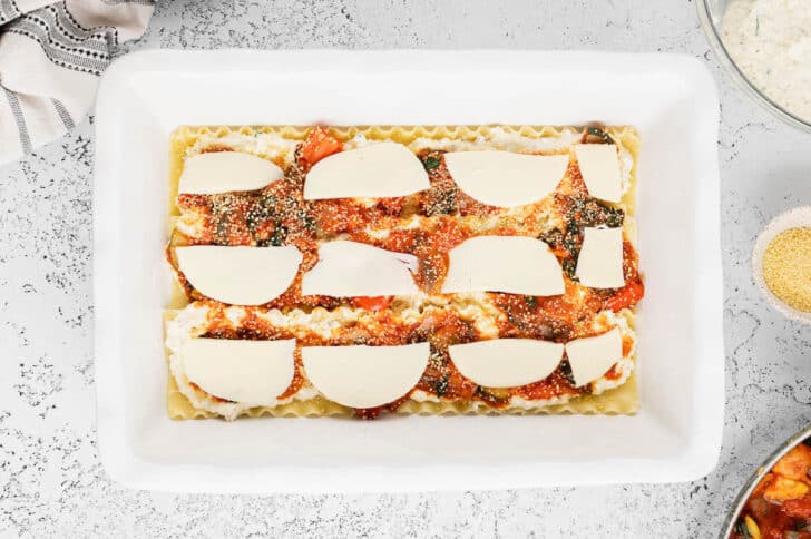 A white lasagna dish layered with lasagna noodles, a cheese mixture, a vegetable marinara mixture, dried couscous and fresh mozzarella half moons.