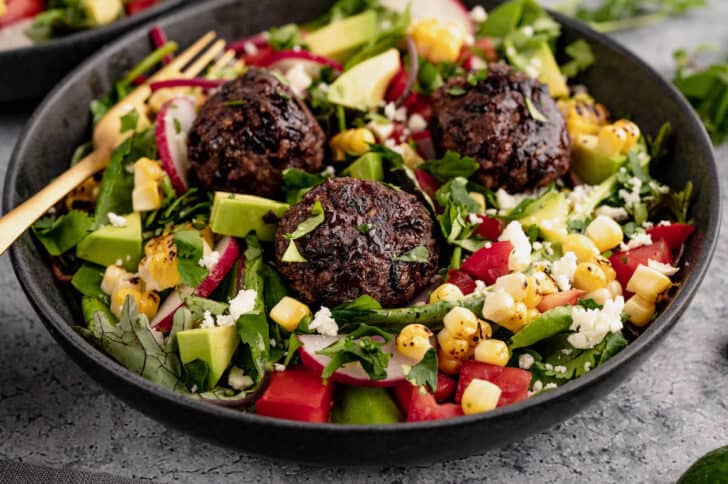 A salad made with greens, chopped tomato, avocado, corn, sliced radishes and cotija cheese topped with black bean balls.