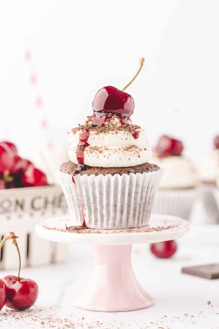A chocolate cupcake topped with whipped cream, chocolate shavings and a cherry, on a small pink cake stand.