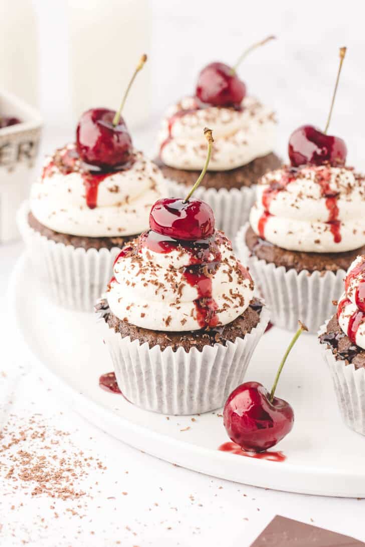 A white plate of Black Forest cupcakes, which are chocolate cupcakes topped with whipped cream, cherries and chocolate shavings.