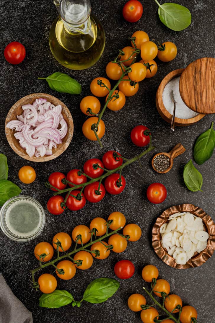 The ingredients for sauteed tomatoes laid out on a dark surface, including cherry tomatoes on their vines, salt and pepper, sliced shallots and garlic, and basil leaves.