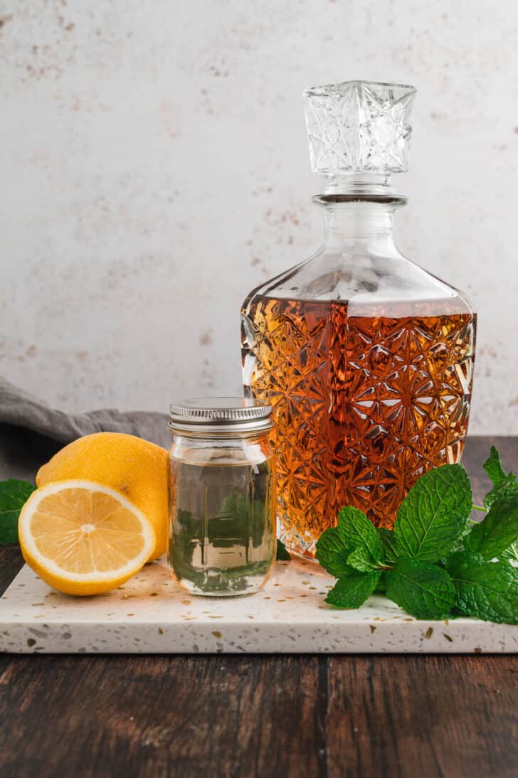 Bourbon smash ingredients including a decorative bottle of whisky, a jar of simple syrup, lemons and fresh mint.