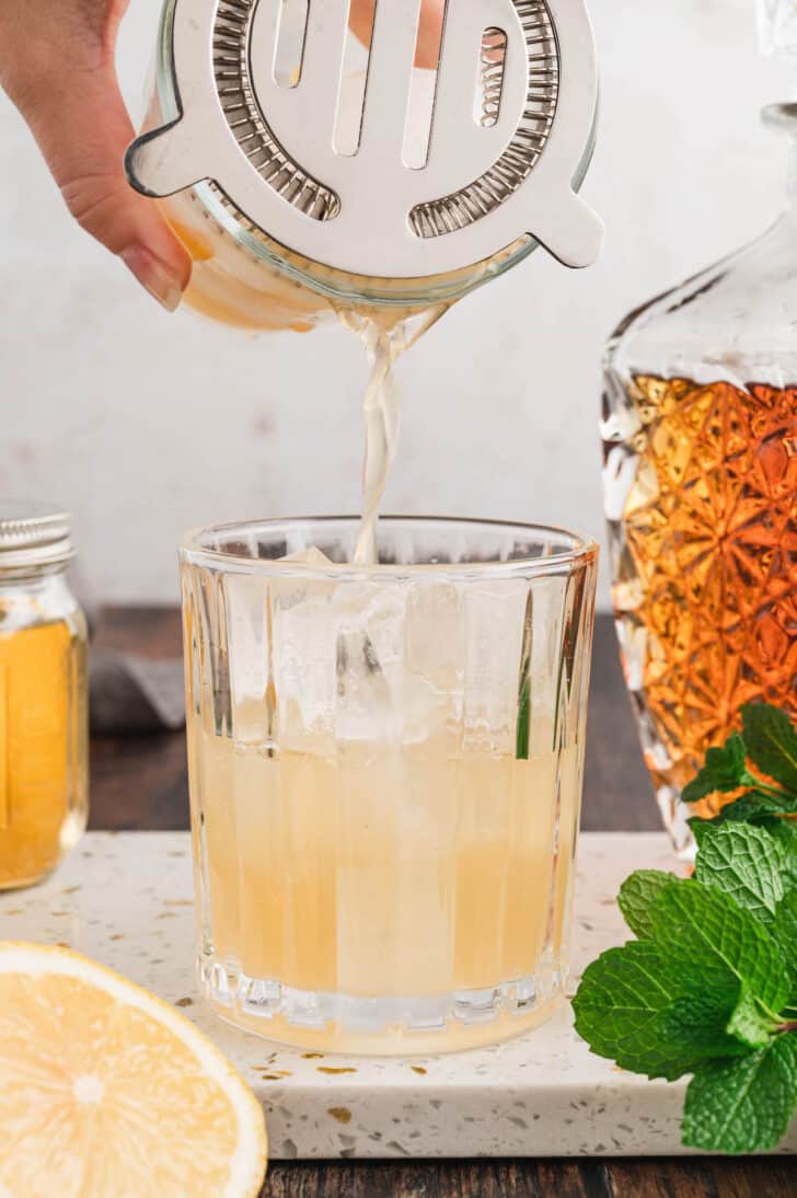 A bourbon smash cocktail being poured from a cocktail shaker with strainer lid into a glass of ice.