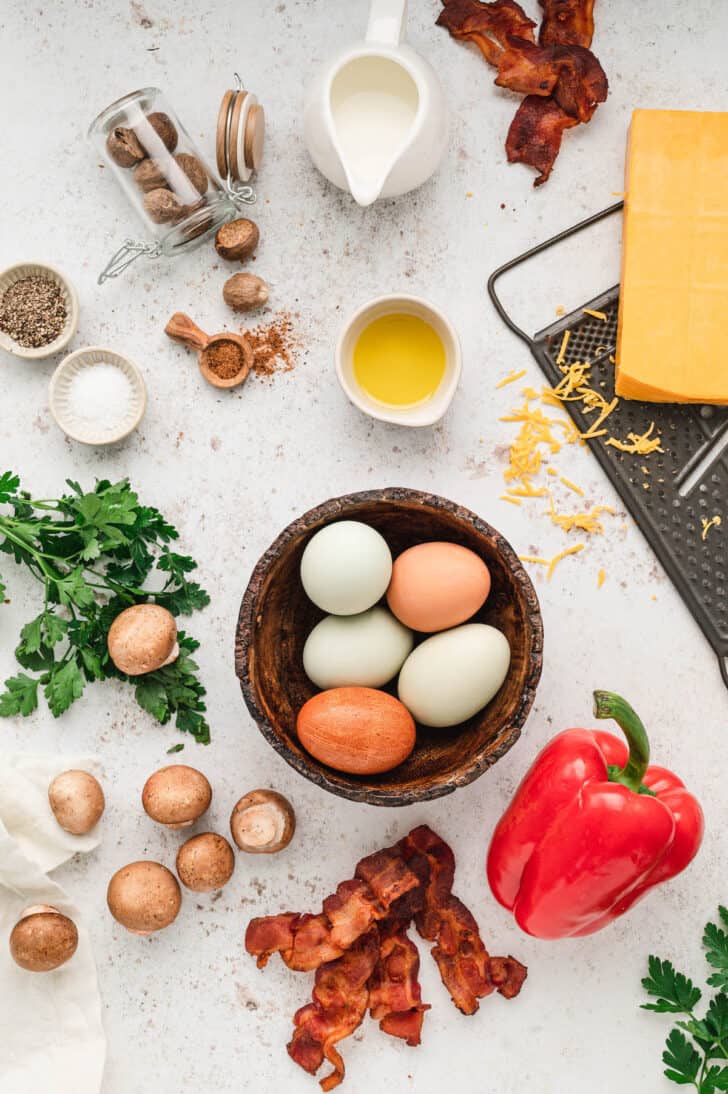 Ingredients laid out on a light surface, including eggs, mushrooms, a red bell pepper, bacon, cheese, herbs and spices.