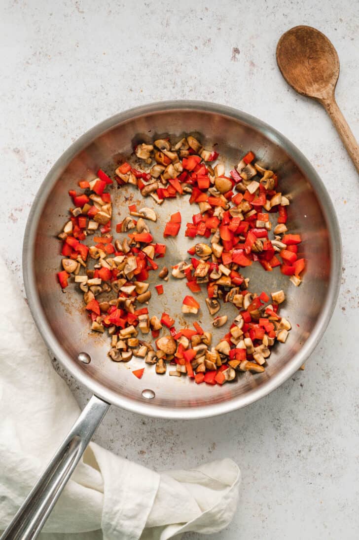 Chopped mushrooms and red bell peppers being sauteed in a stainless steel skillet.