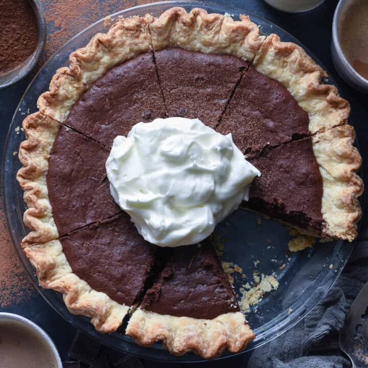 A brownie pie topped with whipped cream, on a table with mugs of coffee and milk.