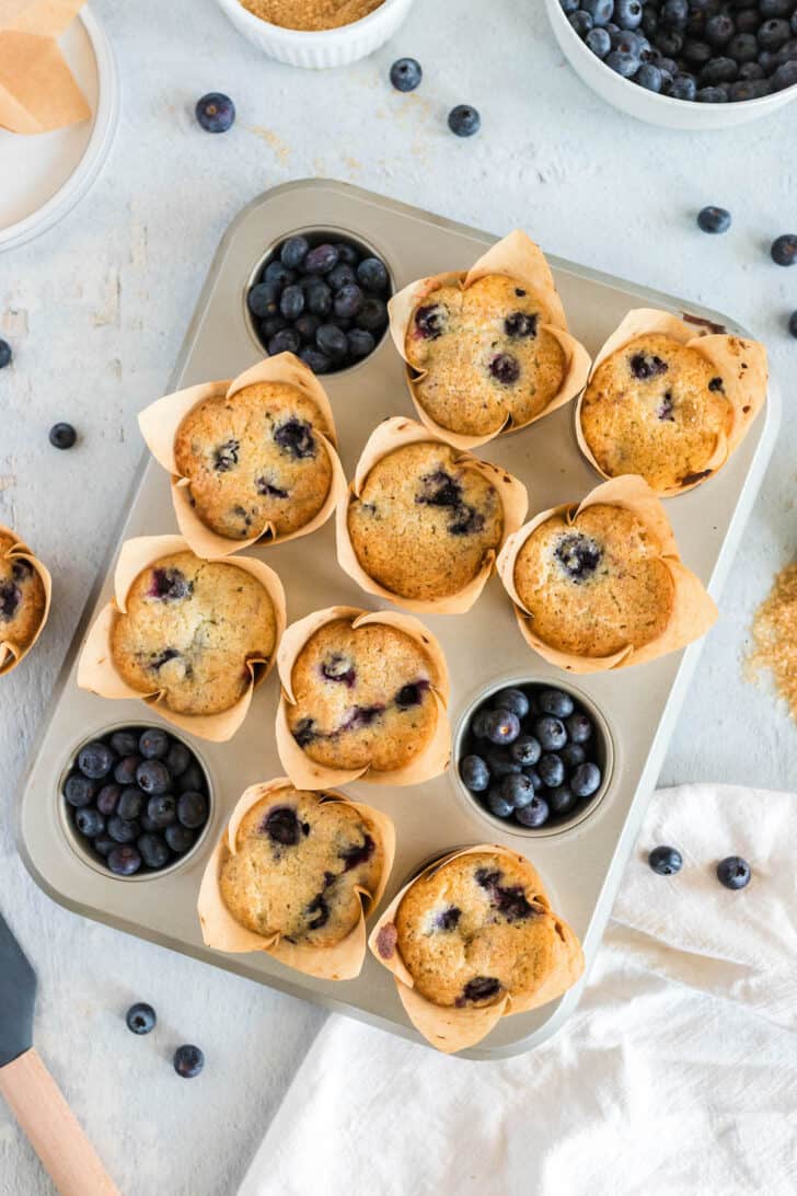 Overhead photo of a muffin pan filled with 9 blueberry muffins. Fresh blueberries fill the other 3 holes.