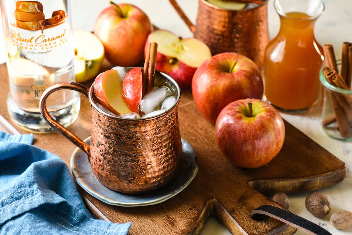 Caramel Apple Pie Moscow Mules - This twist on a classic will take you from summer right into fall. All the best flavors of an apple pie (and caramel!) are added to an ice cold vodka cocktail. | foxeslovelemons.com Wooden cutting board topped with apples and copper mugs filled with apple cider mule cocktails.
