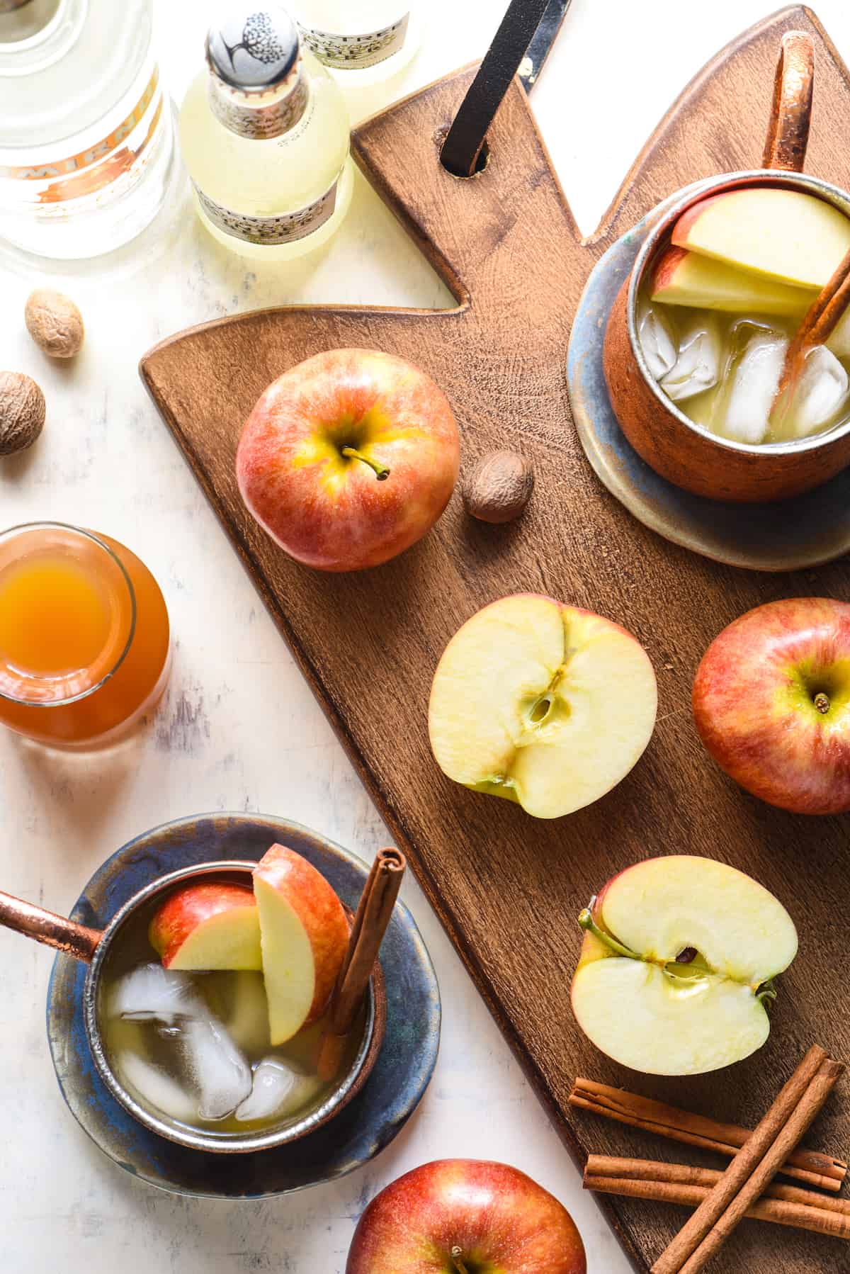 Caramel Apple Pie Moscow Mules - This twist on a classic will take you from summer right into fall. All the best flavors of an apple pie (and caramel!) are added to an ice cold vodka cocktail. | foxeslovelemons.com Overhead shot of a wooden cutting board with cut and whole apples, nutmeg pods, bottles, cinnamon sticks, and copper mugs filled with caramel apple cider mule cocktails.