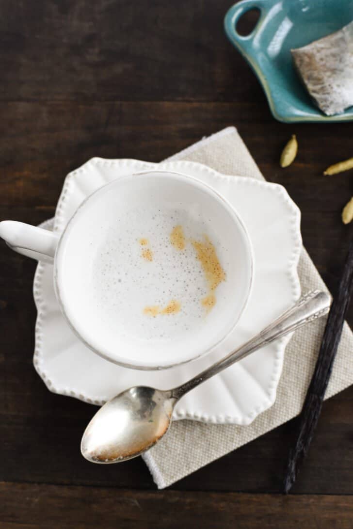 A small white teacup filled with cardamom tea latte on a matching saucer, with a spoon alongside.