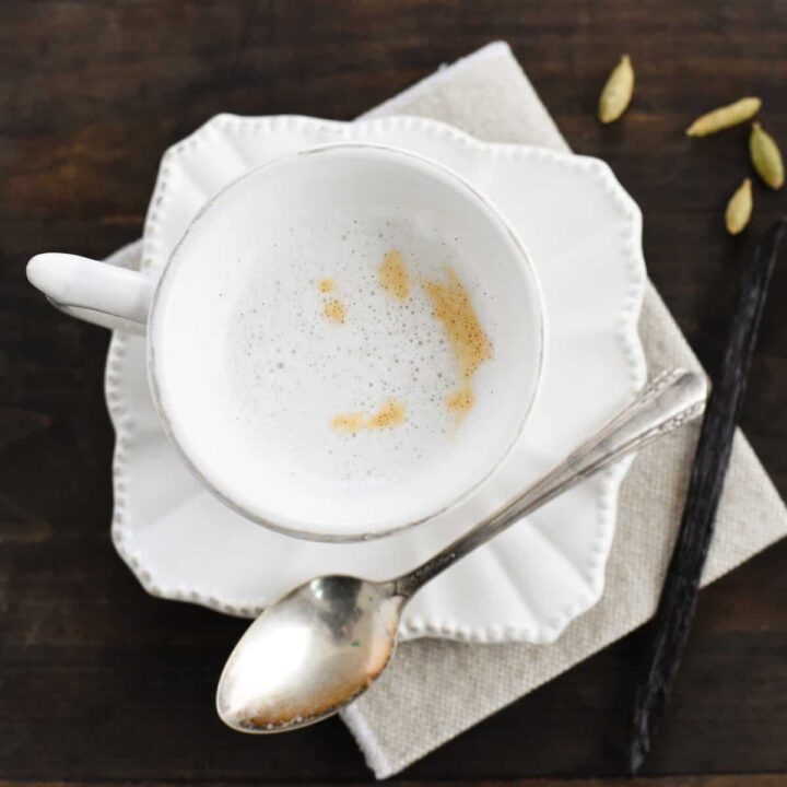 A small white teacup filled with cardamom tea latte on a matching saucer, with a spoon alongside.