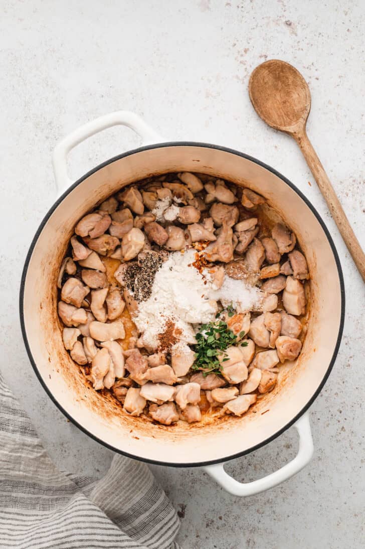 A white Dutch oven filled with browned cubed chicken, flour, spices and herbs.