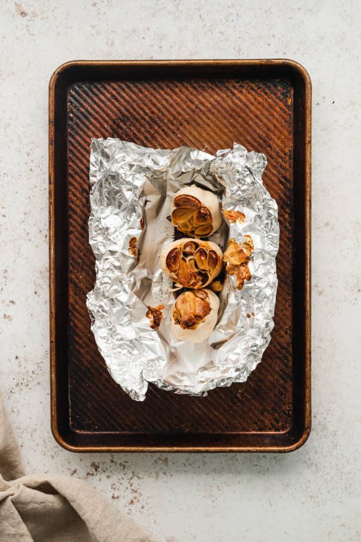 Roasted garlic bulbs in a foil packet on a rimmed baking pan.