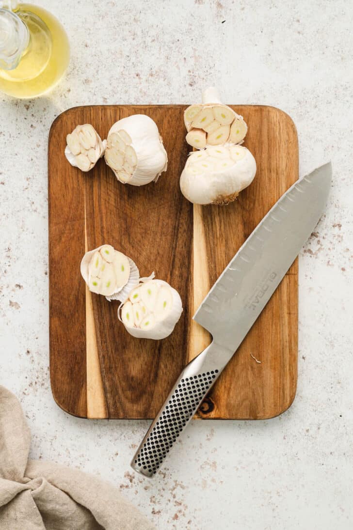 A small wooden cutting board and knife with garlic cloves with their tops sliced off.