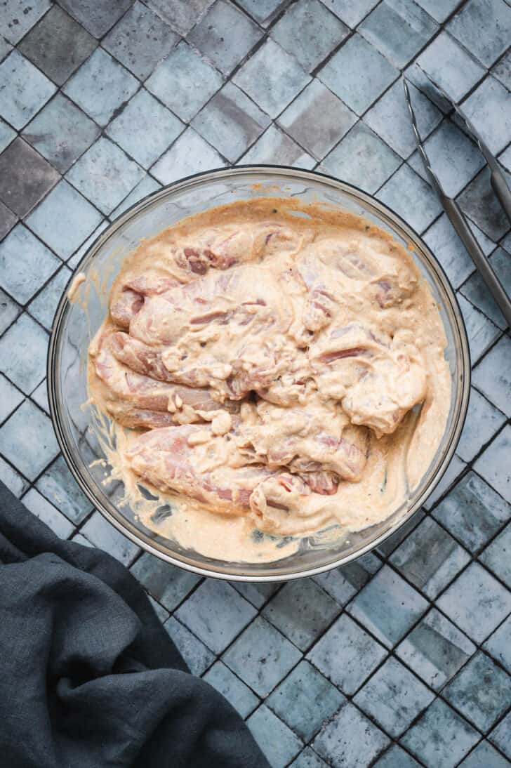 Raw chicken in a orange-hued creamy marinade in a glass bowl.