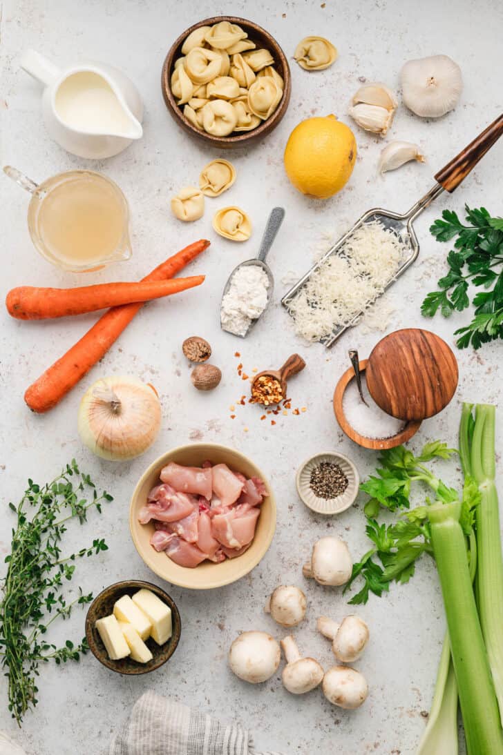 Ingredients laid out on a white surface, including vegtables, herebs, cheese, spices, pasta and chicken.