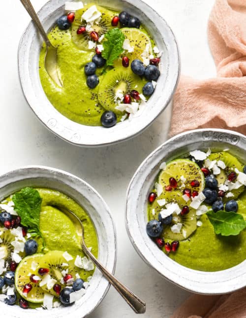 Three fruit smoothie bowls topped with blueberries, pomegranate seeds, coconut, kiwi and a green leaf.