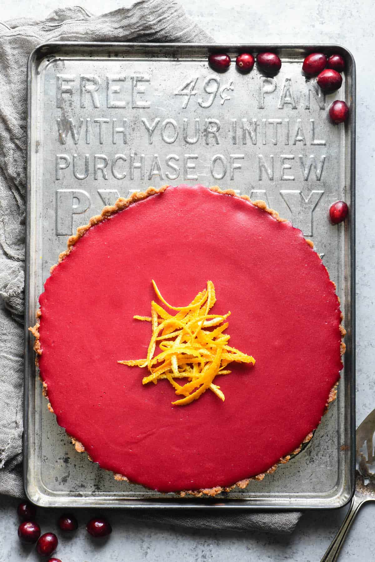 This Cranberry Curd Tart will make a striking addition to your holiday dessert table. Made with fresh cranberries and orange juice, and a graham cracker crust. Can also be made gluten free with an almond or hazelnut crust. | foxeslovelemons.com Whole cranberry tart on vintage baking pan, with fresh cranberries scattered around.