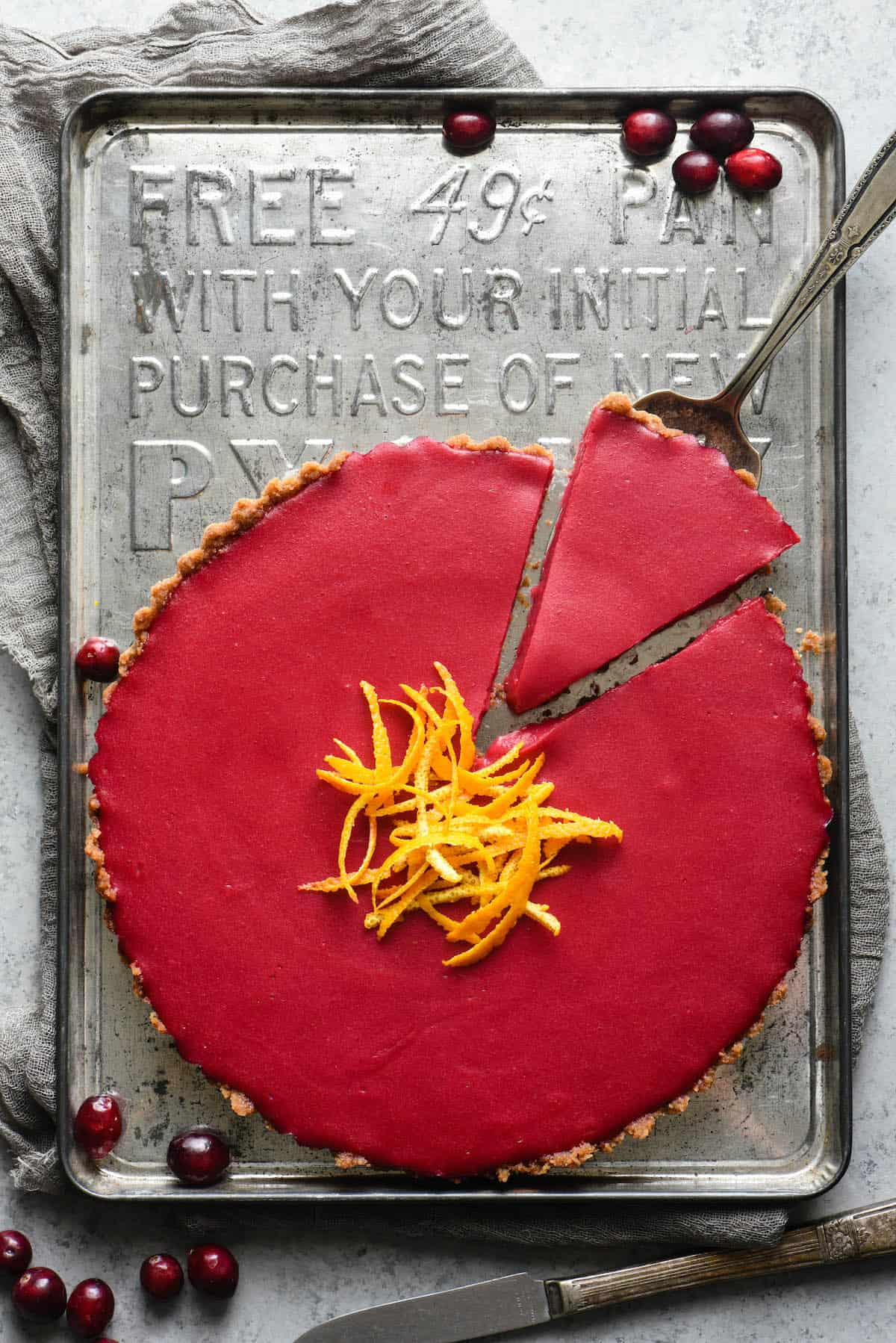 This Cranberry Curd Tart will make a striking addition to your holiday dessert table. Made with fresh cranberries and orange juice, and a graham cracker crust. Can also be made gluten free with an almond or hazelnut crust. | foxeslovelemons.com Magenta hued tart on vintage baking sheet. One piece of tart is being lifted out with a pie server. Fresh cranberries garnish scene.