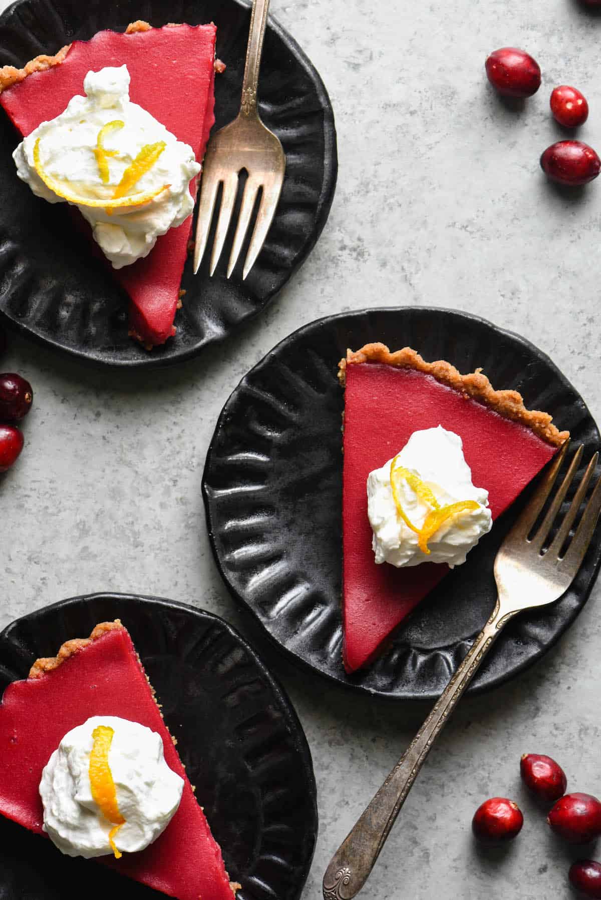 This Cranberry Curd Tart will make a striking addition to your holiday dessert table. Made with fresh cranberries and orange juice, and a graham cracker crust. Can also be made gluten free with an almond or hazelnut crust. | foxeslovelemons.com Overhead photo of three slices of cranberry curd tart on black plates, topped with whipped cream and orange zest.