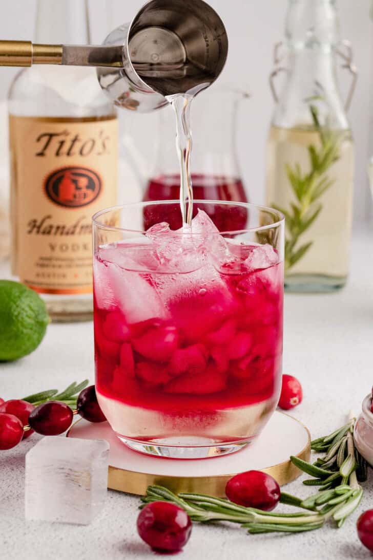 A metal shot glass pouring vodka over a glass filled with ice and red juice.