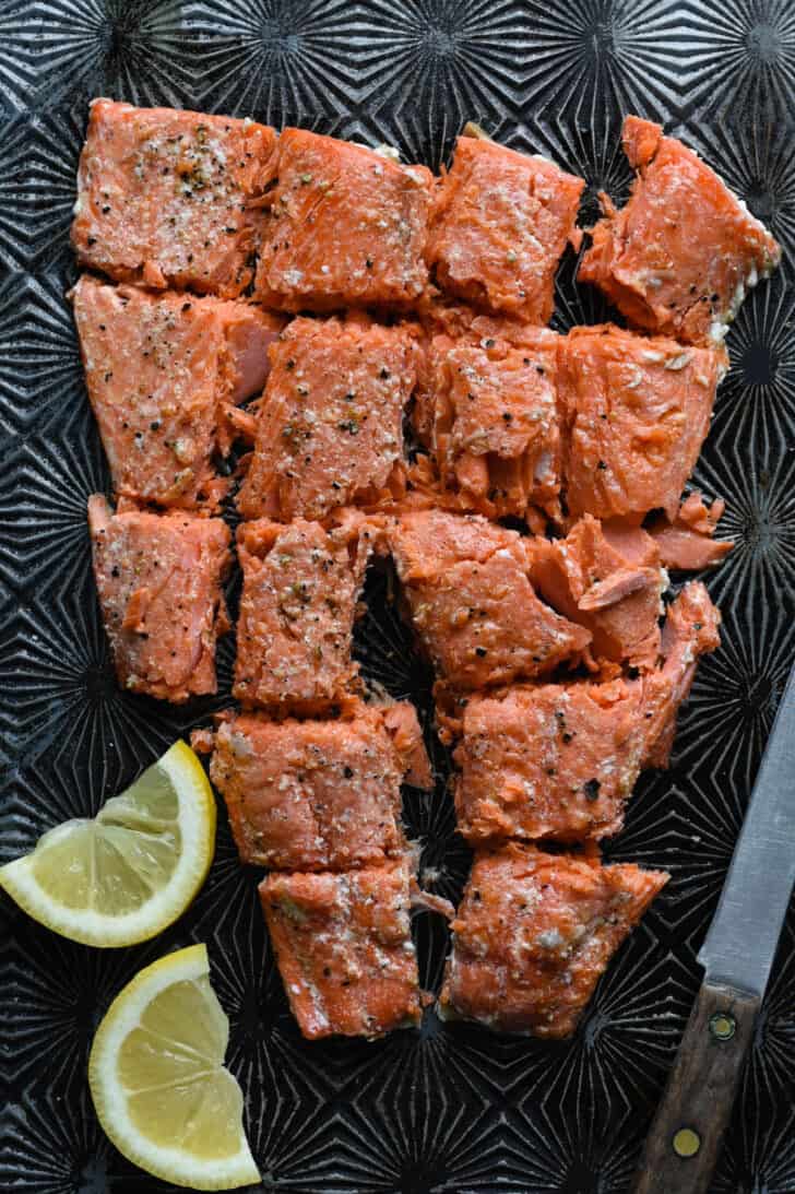 A large fillet of baked orange fish, broken into large chunks, on a textured baking pan with lemon wedges on the side.