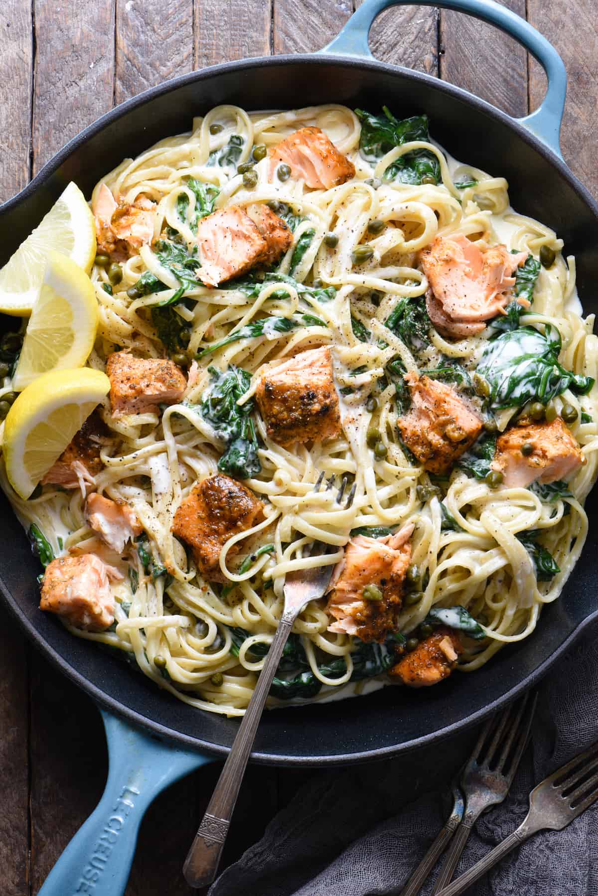 This Creamy Salmon Pasta recipe is a great way to show off what a "home chef" you are. Simple yet impressive, this is a perfect recipe for a date night at home. | foxeslovelemons.com A large blue cast iron skillet filled with creamy salmon pasta with spinach, and lemon wedges as garnish.