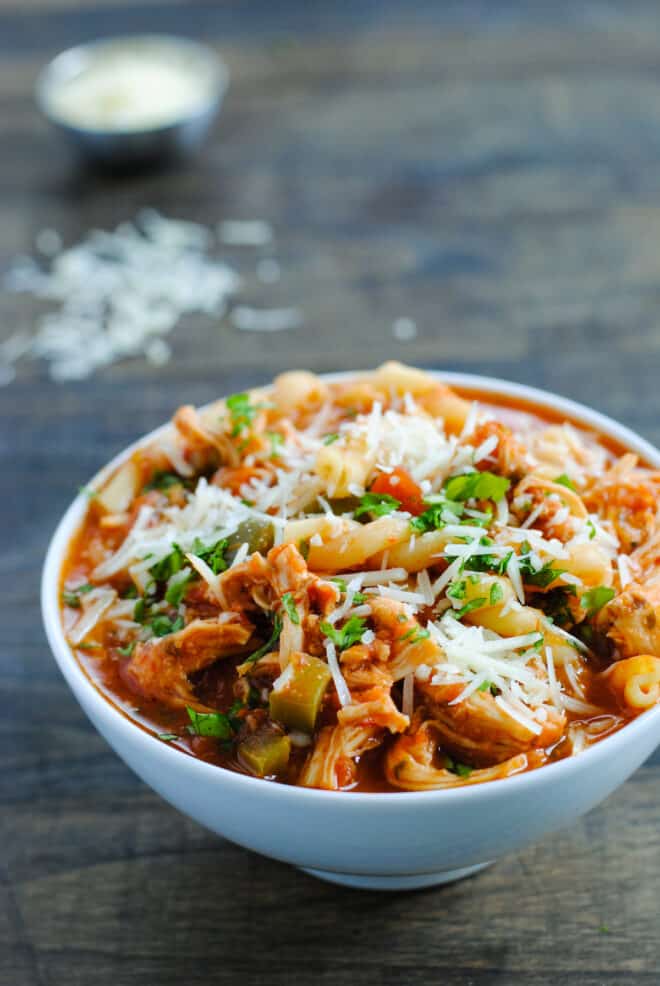 Small white bowl of red broth stew with noodles, vegetables, chicken and cheese.