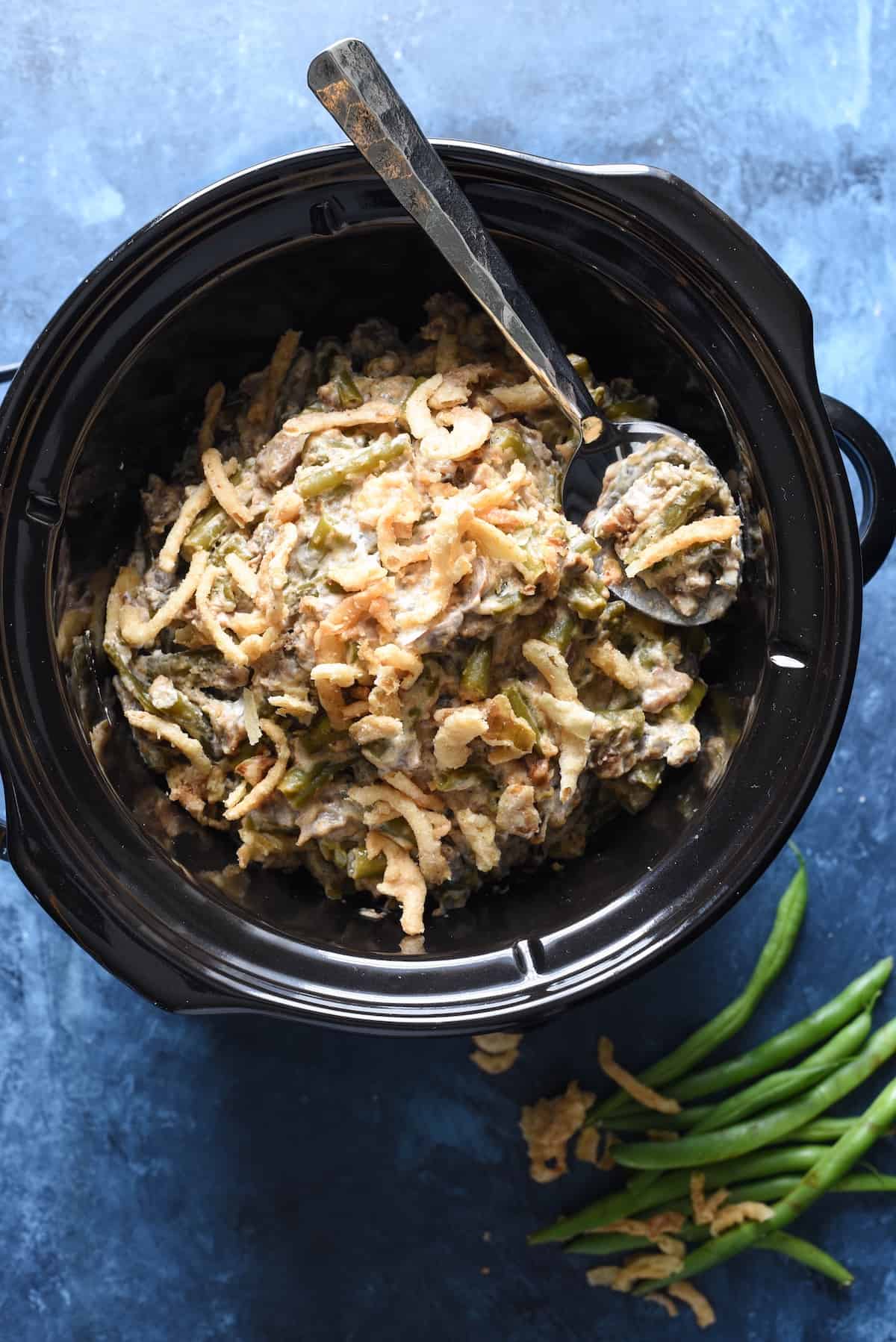 This Crockpot Green Bean Casserole is made with fresh beans and doesn't use canned soup. It's a yummy and easy upgrade to a classic dish, and doesn't take up oven space for a holiday meal! | foxeslovelemons.com A green bean casserole in crock pot on a blue background.