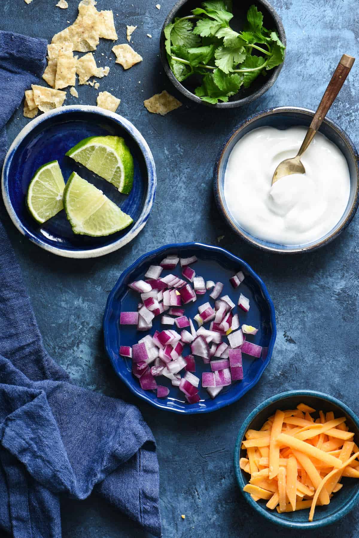 This Crockpot Taco Soup recipe comes together in just a few minutes. Includes instructions for making a large batch and freezing some for another meal, as well as a smaller batch. | foxeslovelemons.com Bowls on blue background filled with garnishes for taco soup crock pot, including cilantro, limes, sour cream, red onion and shredded cheese.