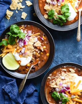 Three black bowl filled with crockpot taco soup and garnished with sour cream, lime, cilantro, cheese, red onion and crushed tortilla chips.