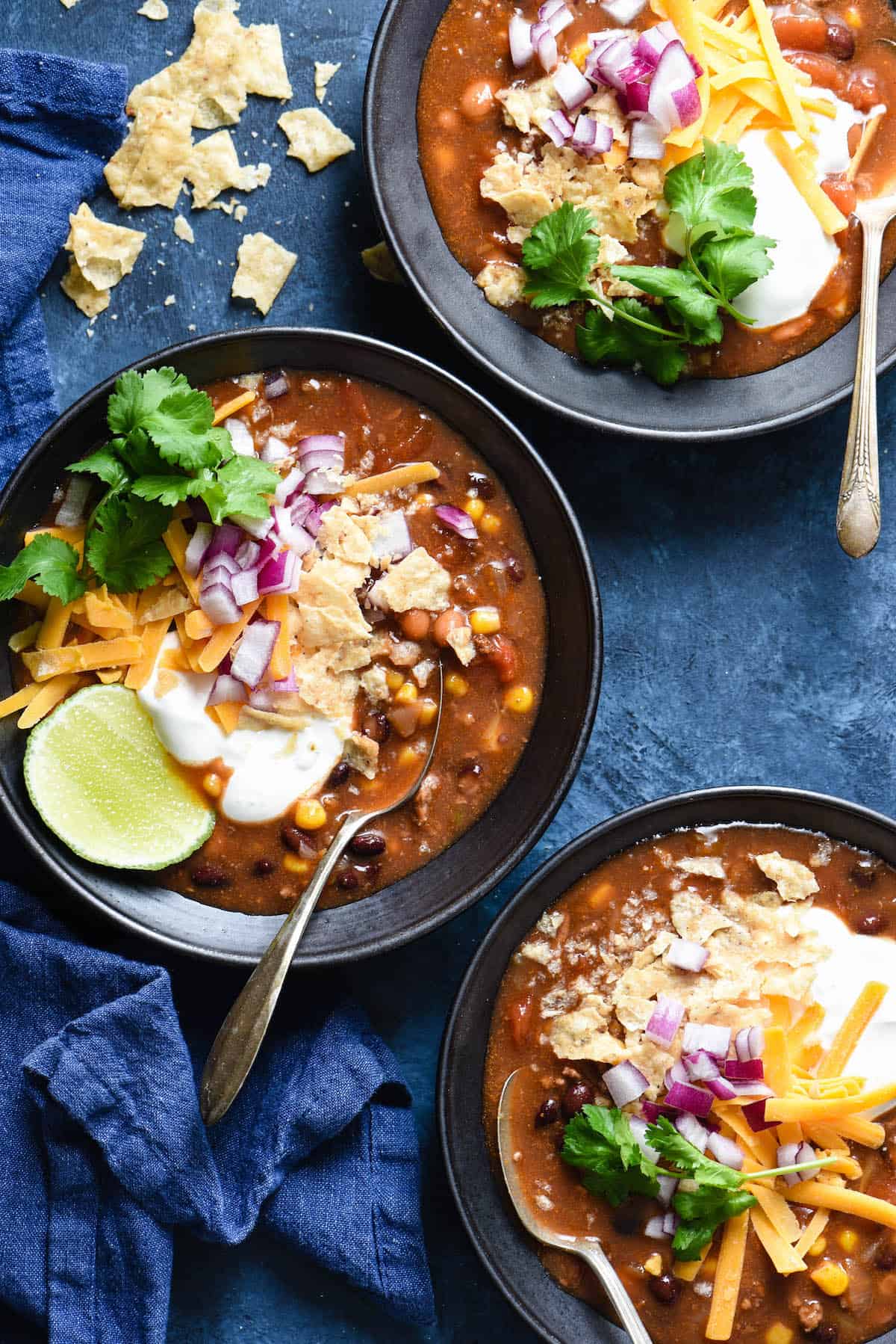 This Crockpot Taco Soup recipe comes together in just a few minutes. Includes instructions for making a large batch and freezing some for another meal, as well as a smaller batch. | foxeslovelemons.com Three black bowls filled with a crockpot taco soup recipe and garnished with sour cream, lime, cilantro, cheese, red onion and crushed tortilla chips.