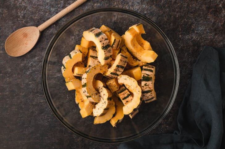 A glass mixing bowl with delicata squash slices being mixed with oil and spices.