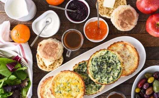 Dark wood tabletop covered with quiche, jam, salad, english muffins, olives and fruit.