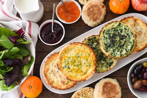 Horizontal overhead shot of brunch buffet table filled with brunch menu ideas like mini quiches, olives, coffee, breakfast breads and spreads, salad and fruit.