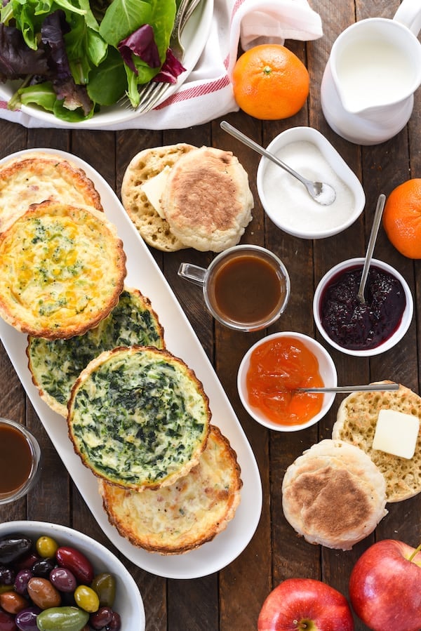 Overhead shot of a brunch buffet table filled with mini quiches, olives, coffee, breakfast breads and spreads, salad and fruit.