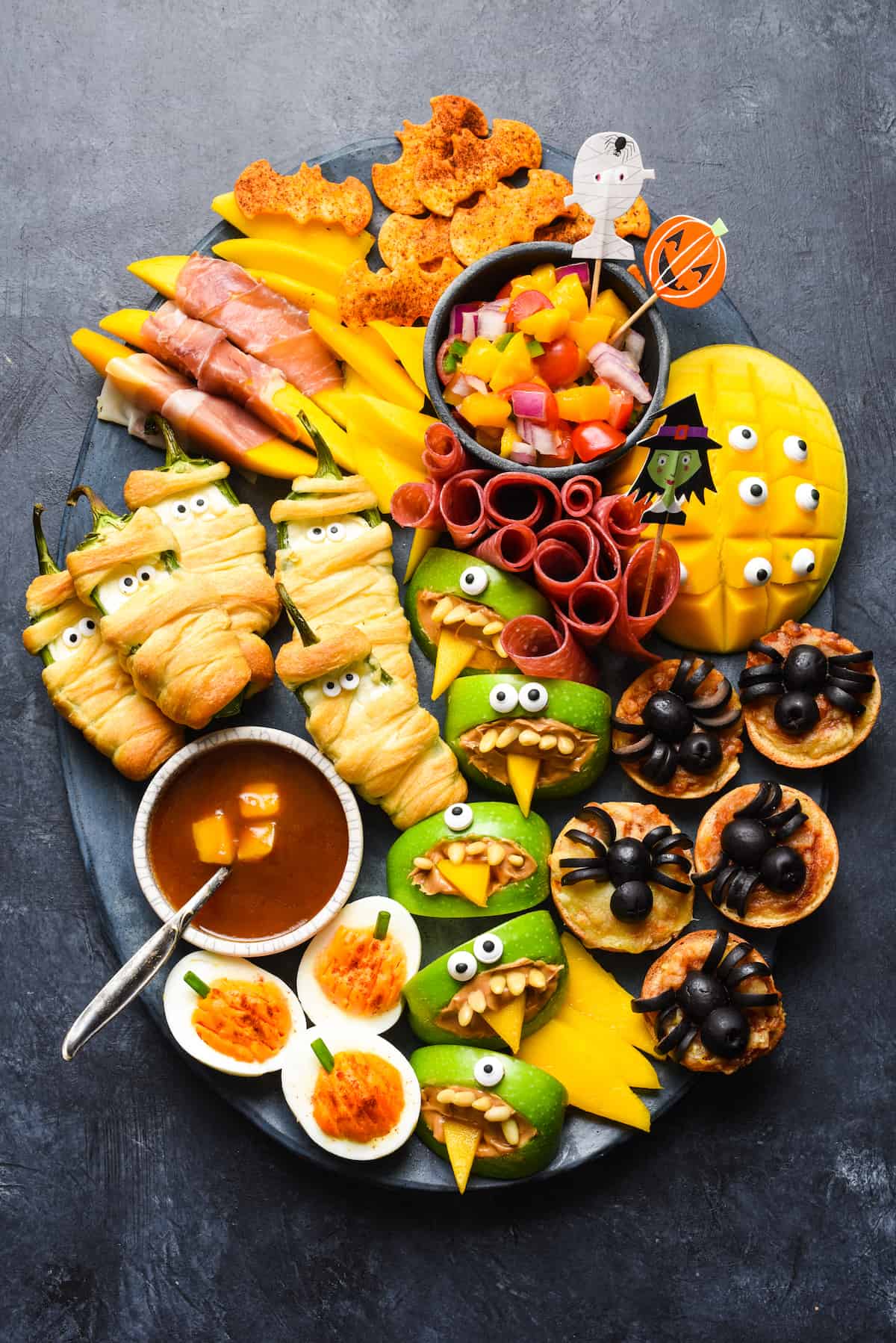 Looking for easy Halloween party food? Look no further than this simple guide to Halloween snacks. Make these edible mummies, spiders and bats for a spooky good time! | foxeslovelemons.com Spooky Halloween food ideas, like jalapeno popper mummies, apple monsters, bat tortilla chips and spider mini pizzas, arranged on a large dark platter on a dark background.