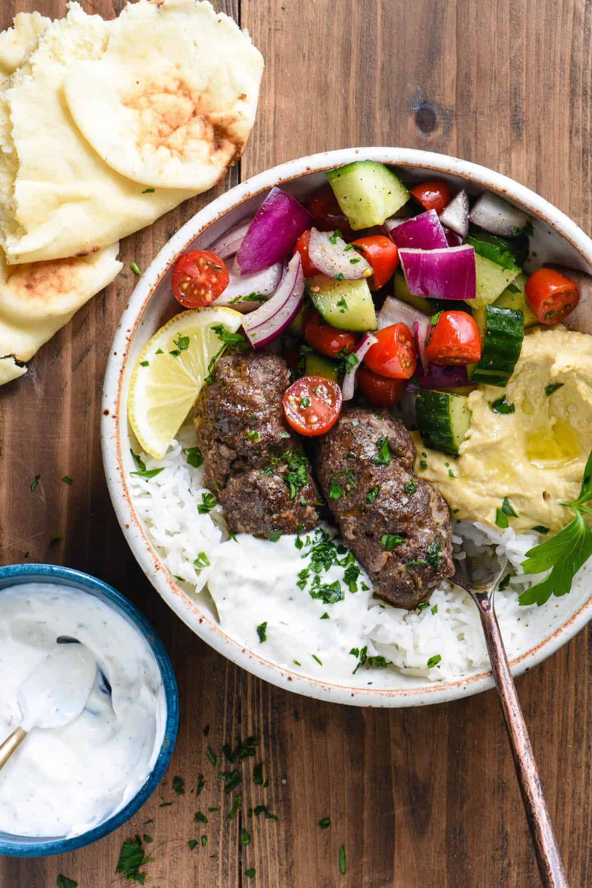 This Easy Kafta Recipe turns a classic Lebanese dish into a simple weeknight meal. This recipe uses ground beef, doesn't make you mess around with skewers, and can be served two different ways - as a sandwich or in a bowl! | foxeslovelemons.com Large bowl filled with rice, kafta, yogurt sauce, hummus and chopped vegetables.