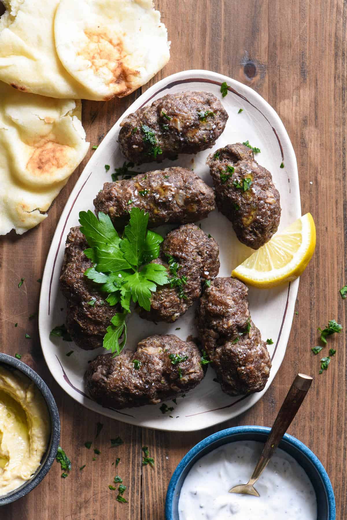 This Easy Kafta Recipe turns a classic Lebanese dish into a simple weeknight meal. This recipe uses ground beef, doesn't make you mess around with skewers, and can be served two different ways - as a sandwich or in a bowl! | foxeslovelemons.com Lebanese beef kafta on small platter garnished with lemon wedge and fresh parsley.