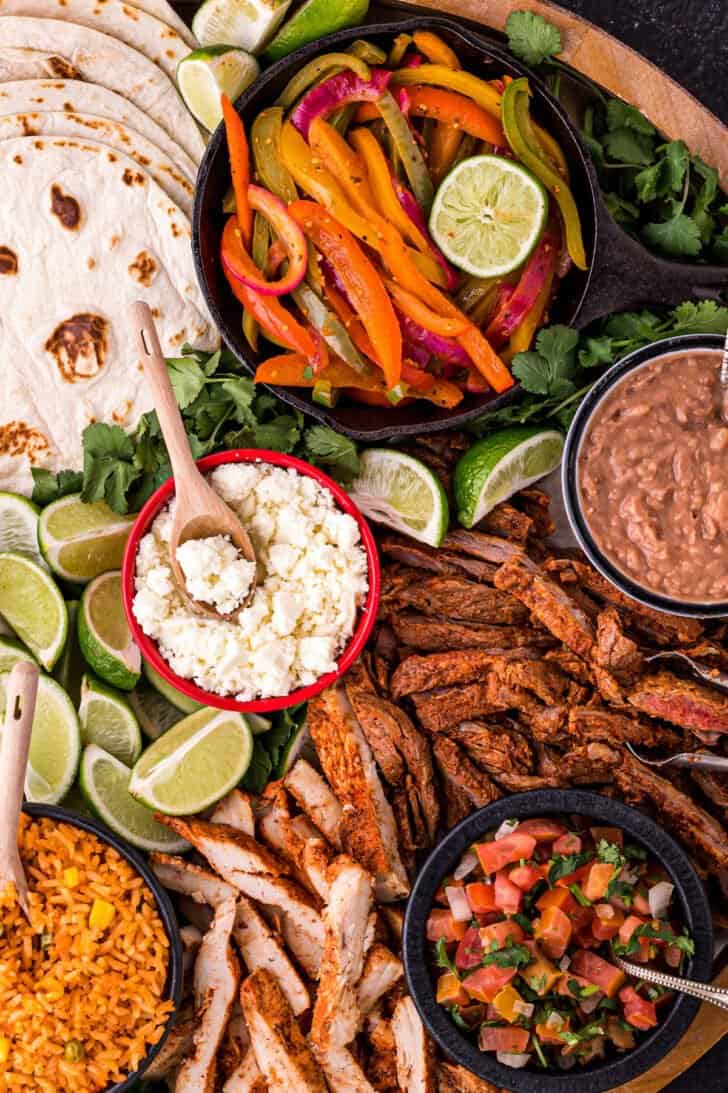 A large round wooden platter filled with fajita bar ingredients like tortillas, cooked chicken and steak, dips, cheese, rice, beans and lime.