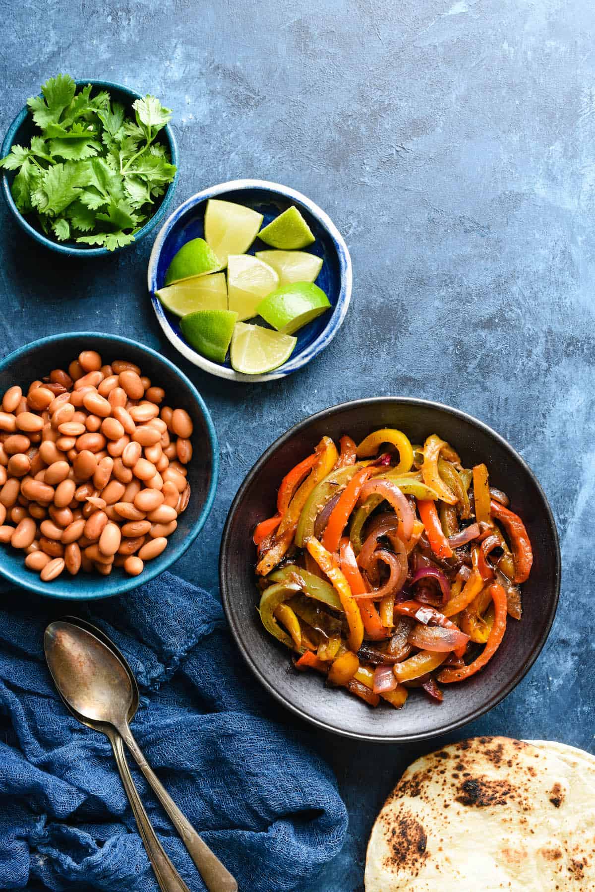 These Fajita Veggies have the restaurant flavor you love, made at home without a seasoning packet. Use them for vegetarian fajitas, or add steak, chicken or shrimp to the mix! | foxeslovelemons.com Bowls of sauteed fajita veggies, pinto beans, lime wedges and cilantro on blue background with charred tortillas.